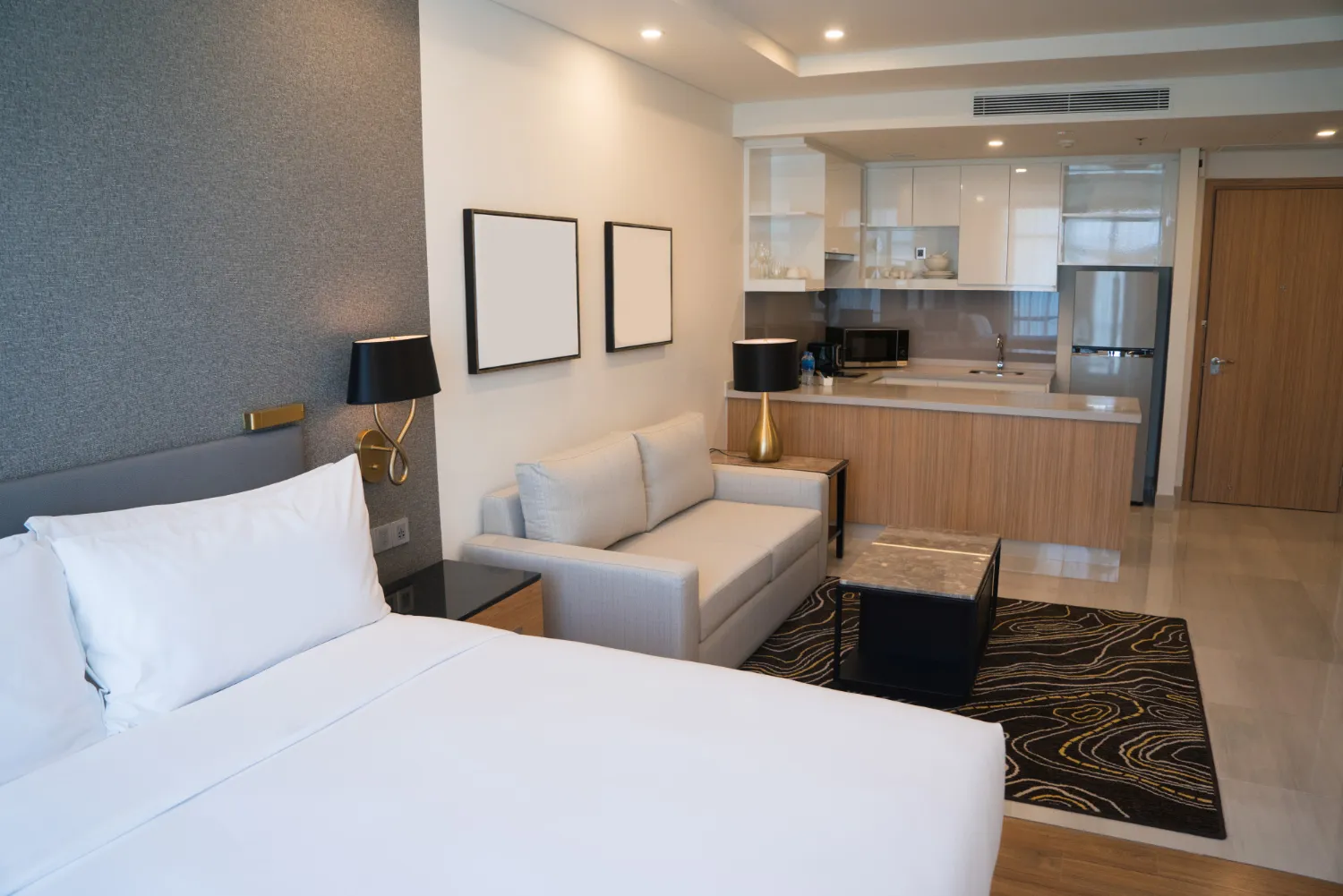 Habitación moderna con cama grande, sofá, mesa de café y cocina equipada, ideal para estancias cómodas y prácticas. Decoración elegante con iluminación suave y detalles en madera.