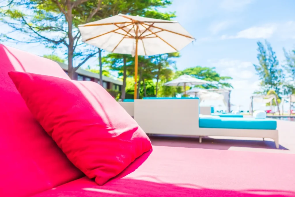 Sofá rojo con cojín en primer plano, rodeado de sombrillas y áreas de descanso al aire libre en un ambiente tropical y soleado. Ideal para relajarse junto a la piscina.