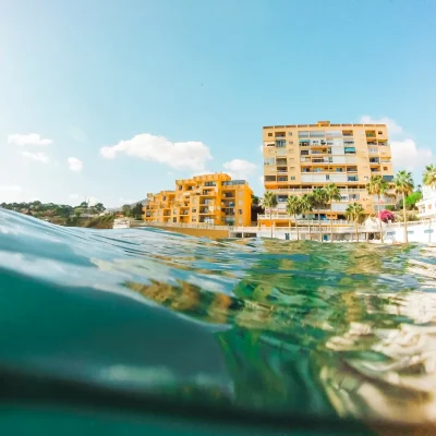 Vista submarina de un complejo residencial amarillo junto a la playa, con palmeras y un cielo despejado. Ideal para promocionar destinos turísticos y actividades acuáticas.